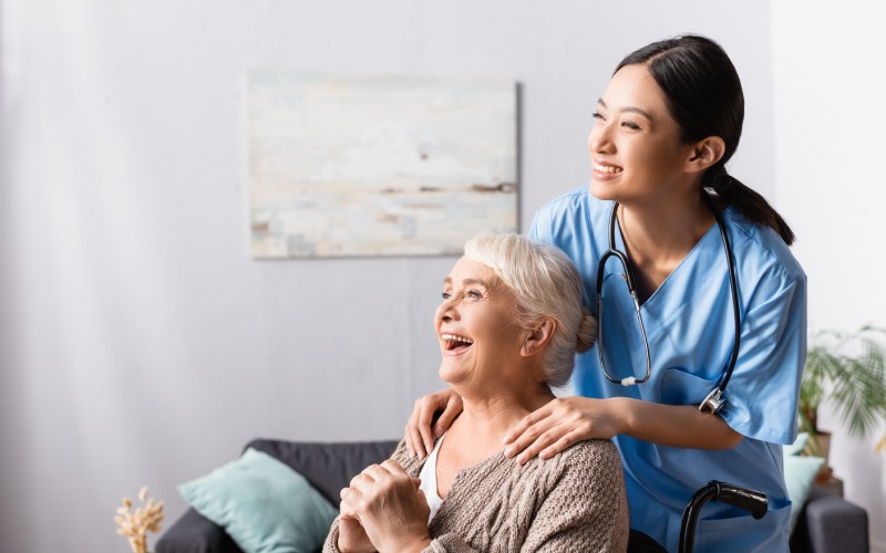happy asian nurse touching shoulders aged handicapped woman laughing wheelchair