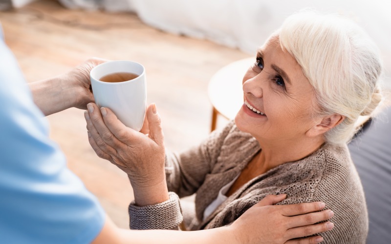 smiling senior woman taking cup tea nurse blurred foreground